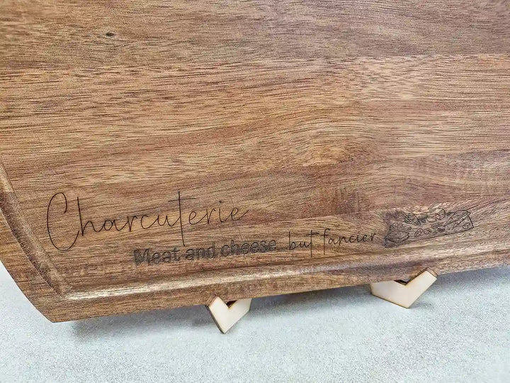 Close-up of the engraved 'Charcuterie. Meat and cheese...but fancier' phrase on the rectangular Acacia wood charcuterie board, highlighting the engraving detail.