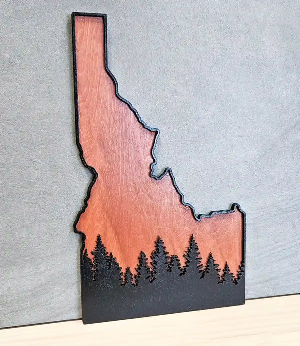 Laser-cut wooden art of the state of Idaho with a forest design backdrop, displayed upright on a light surface. 