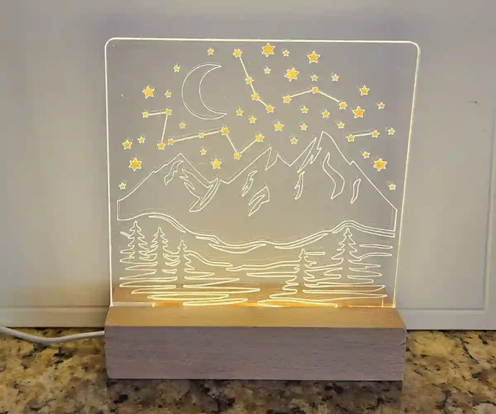 Clear acrylic nightlight with engraved mountain range, crescent moon, stars, and a constellation, illuminated by a light base.
