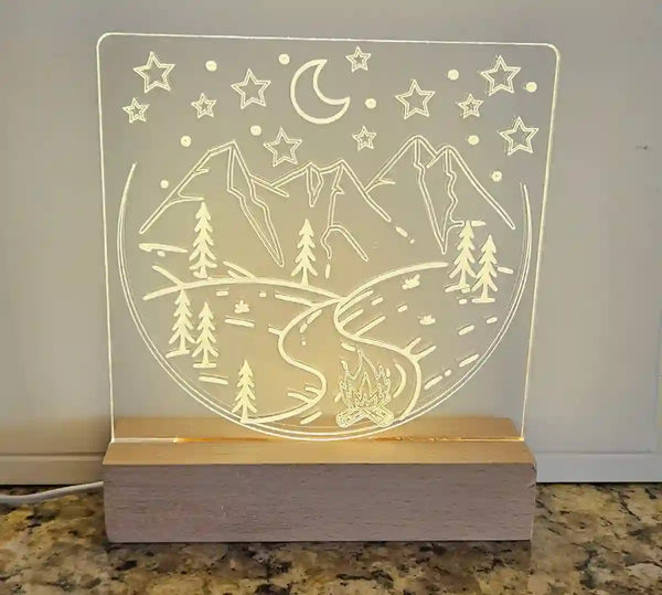 Clear acrylic nightlight with engraved mountain landscape, crescent moon, stars, and a campfire, illuminated by a light base.