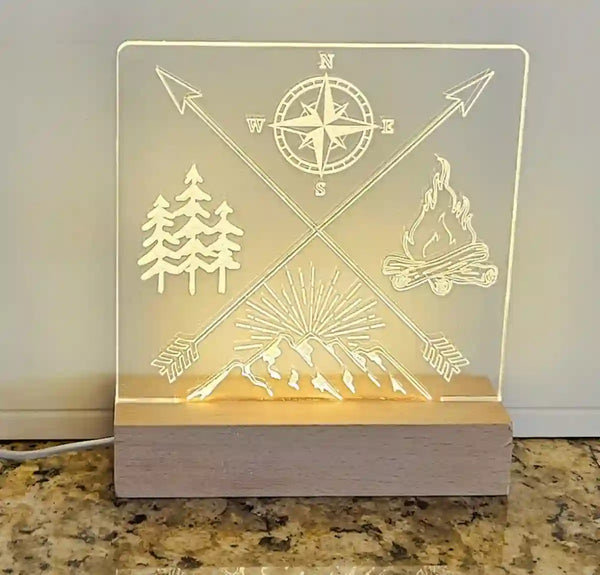 Clear acrylic nightlight with engraved crossed arrows, compass, mountain range, pine trees, and campfire, illuminated by a light base.