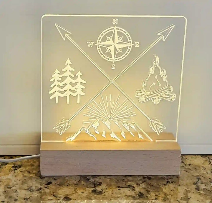 Clear acrylic nightlight with engraved crossed arrows, compass, mountain range, pine trees, and campfire, illuminated by a light base.