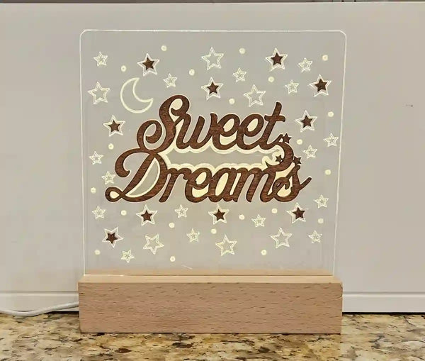 Illuminated nightlight with a clear acrylic panel featuring 'Sweet Dreams' in script, a crescent moon, and stars, resting on a wooden base, against a light countertop and white background.