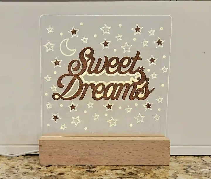 Illuminated nightlight with a clear acrylic panel featuring 'Sweet Dreams' in script, a crescent moon, and stars, resting on a wooden base, against a light countertop and white background.