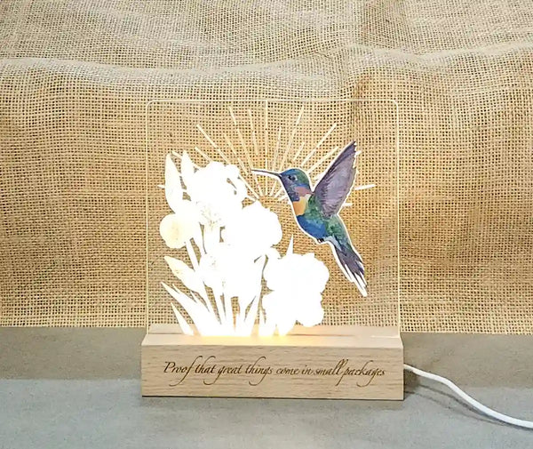 Clear acrylic nightlight with layered and engraved hand-painted hummingbird and engraved iris design, illuminated by a light base, with a wooden base featuring the engraved phrase 'Proof that great things come in small packages'.