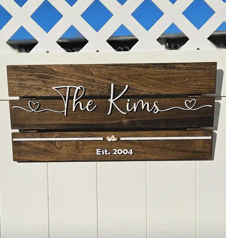 Large rectangular wooden family name sign with 'The Kims' in white script font, two hearts, and 'Est. 2004' in white block font, against a white lattice background.