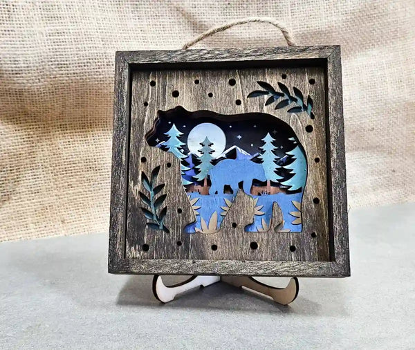 Front-facing view of the square wooden shadow box with a bear silhouette against a moonlit forest scene, displayed on a stand against a burlap background.