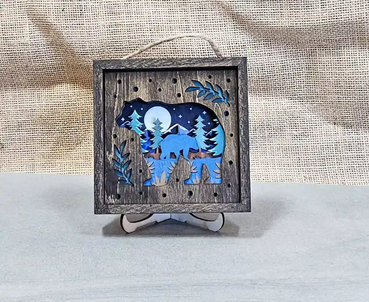 Square wooden shadow box with layered design, featuring a bear silhouette against a moonlit forest scene, displayed on a stand against a burlap background.