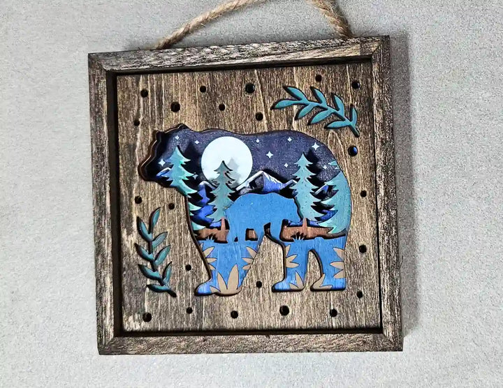 Close-up of the layered bear and moon design within a square wooden shadow box, highlighting the intricate cutouts and depth, against a textured gray background.