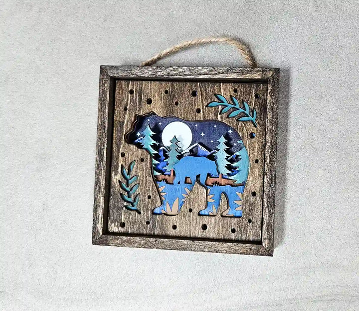 Angled view of the square wooden shadow box with a bear and moon design, showing the layered wood effect and a rope hanger, against a gray background.