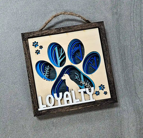 Angled close-up of the square wooden shadow box, showing the layered paper and wood dog paw print design, dog silhouette, and 'Loyalty' text, against a gray background.