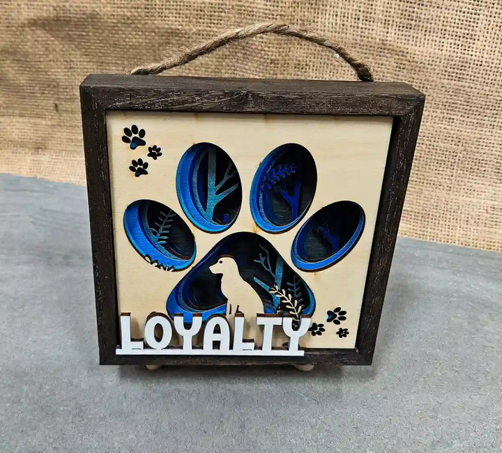 Square wooden shadow box with layered paper and wood design featuring a dog paw print, dog silhouette, and the word 'Loyalty,' displayed on a textured gray and wood surface with a burlap backdrop.