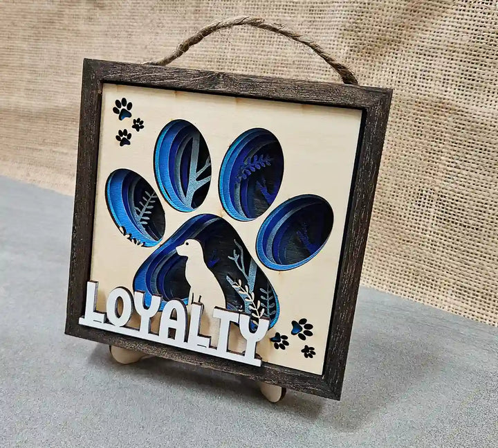 Square wooden shadow box with layered paper and wood design featuring a dog paw print, dog silhouette, and the word 'Loyalty,' displayed on a textured gray and wood surface with a burlap backdrop and stand.