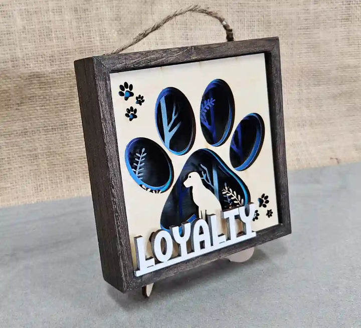 Angled view of the square wooden shadow box, highlighting the layered paper and wood dog paw print design, dog silhouette, and 'Loyalty' text, against a gray and burlap background.