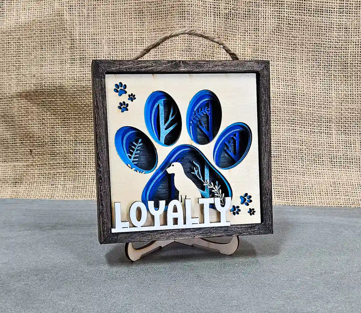 Front view of the square wooden shadow box with layered paper and wood design featuring a dog paw print, dog silhouette, and the word 'Loyalty,' displayed on a stand against a gray and burlap background.