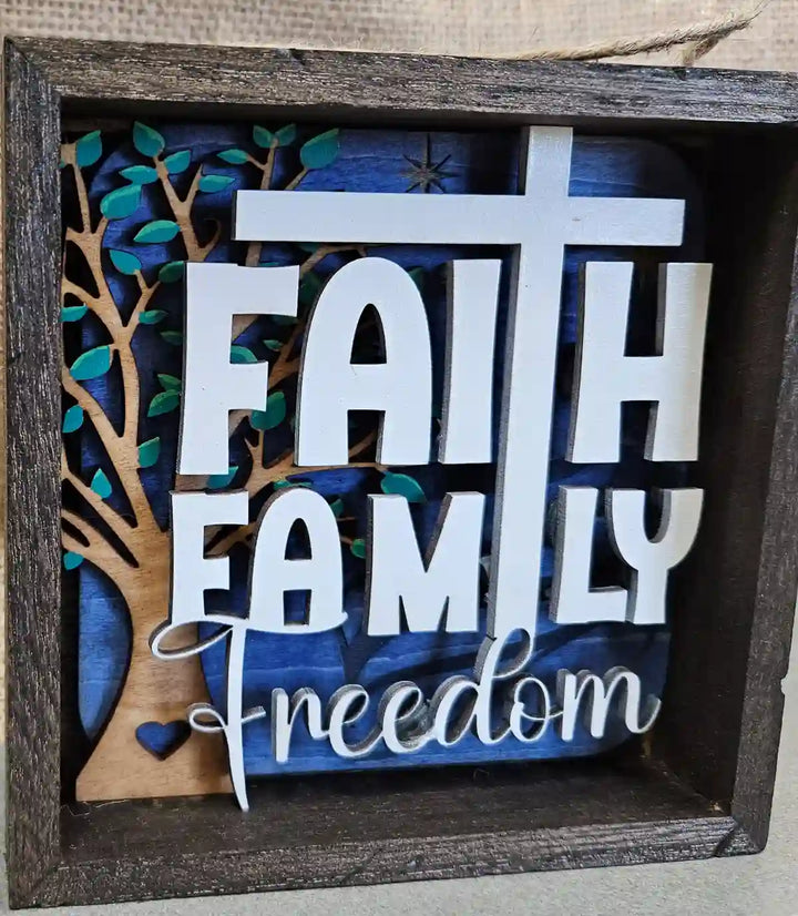 Square wooden shadow box with layered design, featuring the words 'Faith Family Freedom' in white lettering, a white cross, a tree silhouette, and a small heart, against a blue and wood background.