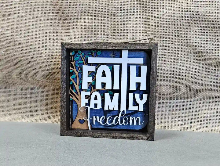 Square wooden shadow box with layered design, displaying the words 'Faith Family Freedom,' a cross, a tree silhouette, and a heart, against a blue and wood background, with a burlap backdrop.