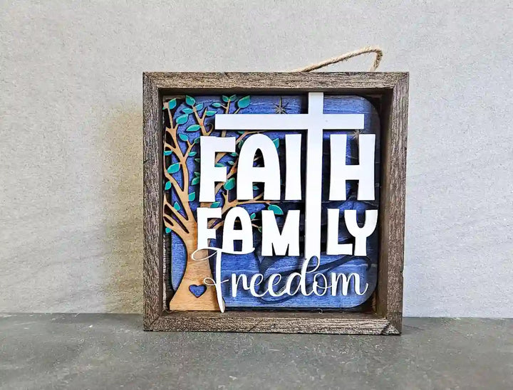 Square wooden shadow box with layered design, featuring 'Faith Family Freedom' text, a cross, a tree silhouette, and a heart, against a blue and wood background, displayed on a textured gray surface.