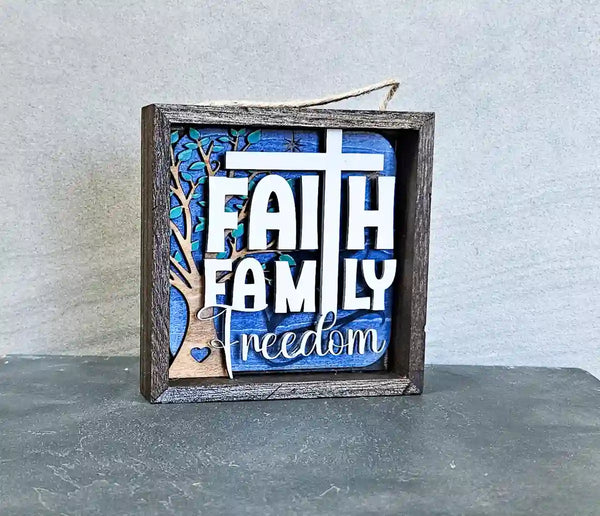 Angled view of the square wooden shadow box, highlighting the layered design with 'Faith Family Freedom' text, a cross, a tree, and a heart, against a gray surface.
