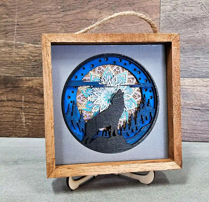 Square wooden shadow box with layered mandala and wolf design, featuring a howling wolf silhouette against a mandala pattern, displayed on a stand with a gray background.