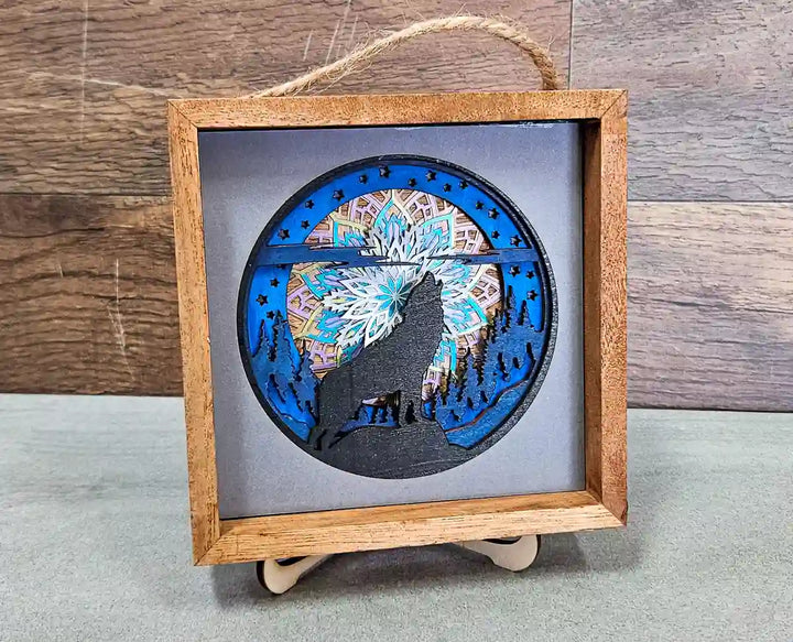 Angled view of the square wooden shadow box, showing the layered mandala and wolf design, with a rope hanger, against a gray background.
