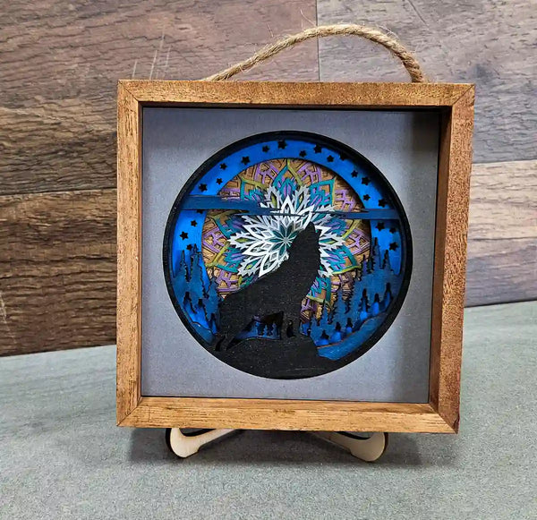 Square wooden shadow box with layered mandala and wolf design, displayed on a stand, against a gray and wood background.