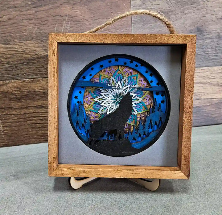 Square wooden shadow box with layered mandala and wolf design, displayed on a stand, against a gray and wood background.