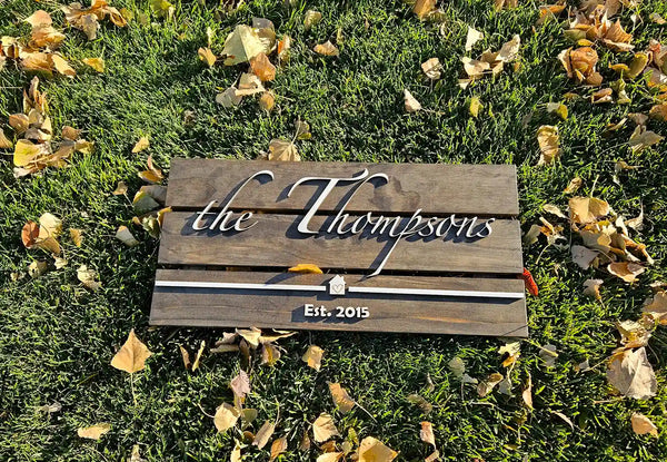 Large rectangular wooden family name sign with 'the Thompsons' in white script font, a small house shape, and 'Est. 2015' in white block font, laying on grass with fallen leaves.