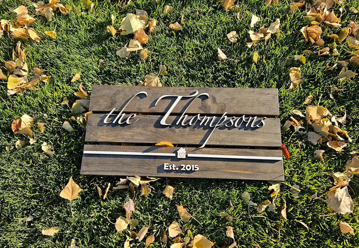 Large rectangular wooden family name sign with 'the Thompsons' in white script font, a small house shape, and 'Est. 2015' in white block font, laying on grass with fallen leaves.