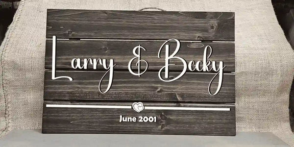 Medium rectangular personalized wooden sign with 'Larry & Becky' in white script font, a small dual heart shape, and 'June 2001' in white block font, against a burlap background.