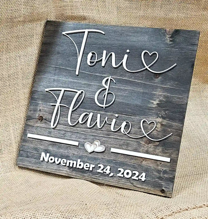 Small square wooden sign with 'Tonie & Flavio' in white script font, two hearts, and 'November 24, 2024' in white block font, displayed at an angle against a burlap background.