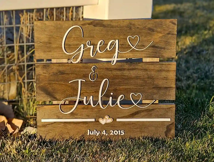 Medium square wooden sign with 'Greg & Julie' in white script font, two hearts, and 'July 4, 2015' in white block font, displayed outdoors on grass.