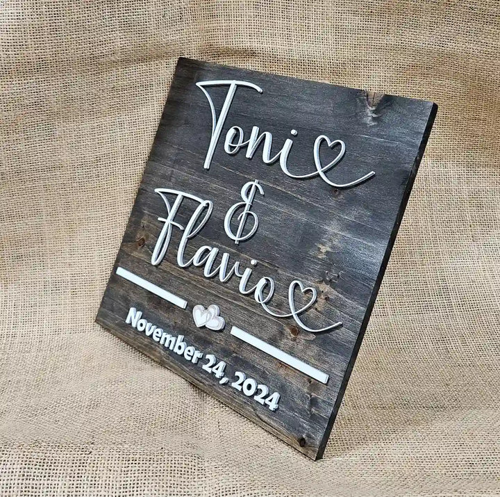 Angled close-up of a small square wooden sign with 'Tonie & Flavio' in white script font, two hearts, and 'November 24, 2024' in white block font, against a burlap background.