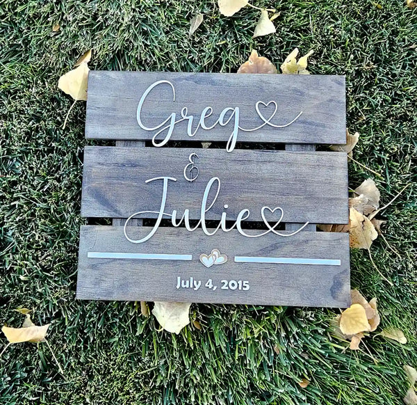 Medium square wooden sign with 'Greg & Julie' in white script font, two hearts, and 'July 4, 2015' in white block font, from a top-down perspective on grass with leaves.