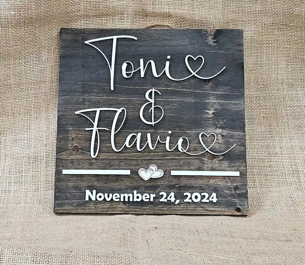Small square wooden sign with 'Tonie & Flavio' in white script font, two hearts, and 'November 24, 2024' in white block font, laying flat against a burlap background.