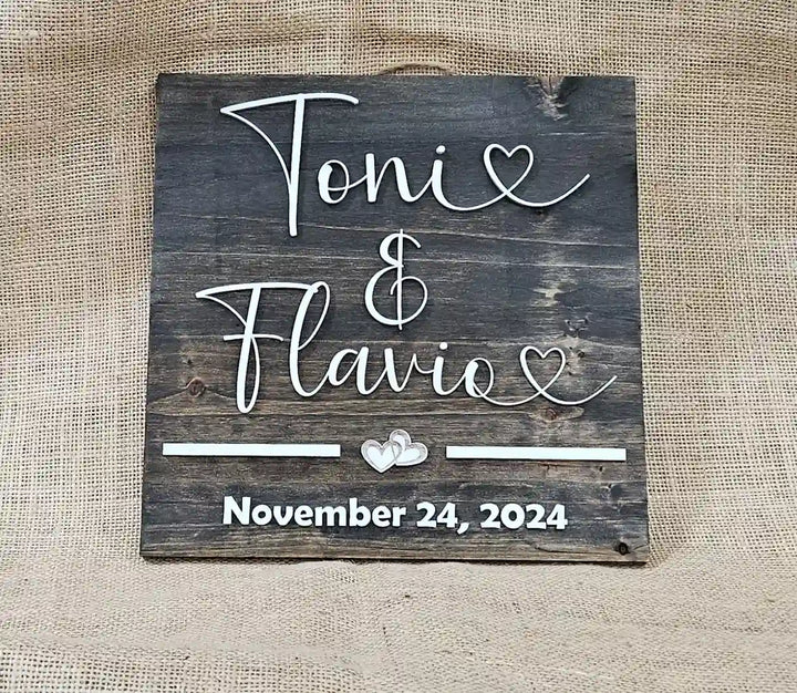 Small square wooden sign with 'Tonie & Flavio' in white script font, two hearts, and 'November 24, 2024' in white block font, laying flat against a burlap background.