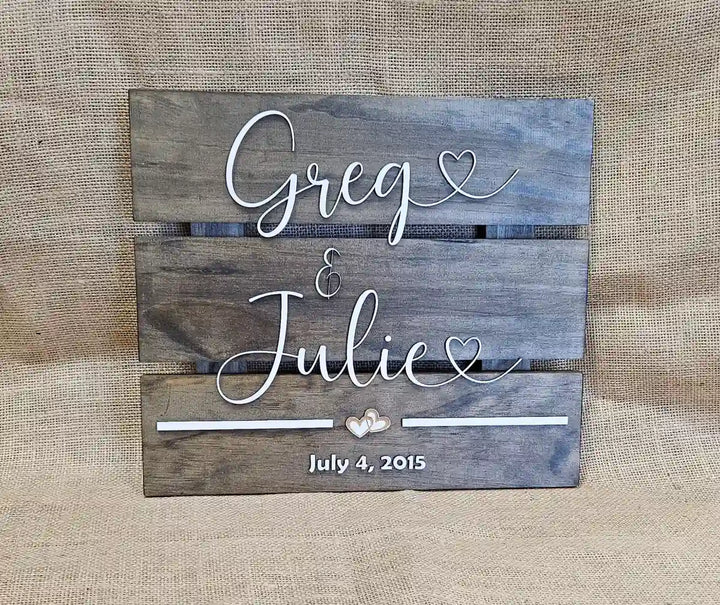 Medium square wooden sign with 'Greg & Julie' in white script font, two hearts, and 'July 4, 2015' in white block font, against a burlap background.