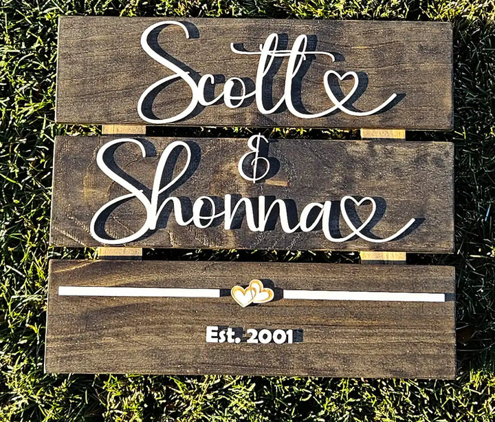 Medium square wooden sign with 'Scott & Shonna' in white script font, two hearts, and 'Est. 2001' in white block font, from a top-down perspective on grass.