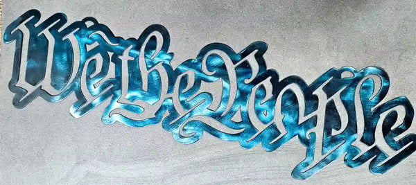 Close-up of a steel sign cut out in the script lettering of 'We the People' with a blue and silver variegated finish, lying on a textured gray surface.