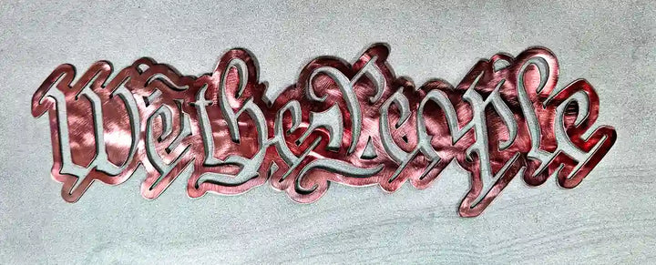 Close-up of a steel sign cut out in the script lettering of 'We the People' with a Cranberry Glass finish, lying on a textured gray surface.