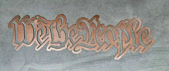 Close-up of a steel sign cut out in the script lettering of 'We the People' with a Rose Gold finish, lying on a textured gray surface.