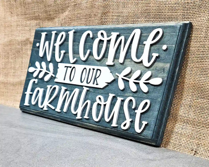 Angled view of a rectangular wooden sign with a blue stained finish and raised white script lettering that reads 'Welcome' at the top and 'farmhouse' at the bottom, with a white arrow pointing right that says 'TO OUR' in smaller black letters, flanked by white leafy branches, against a burlap background.