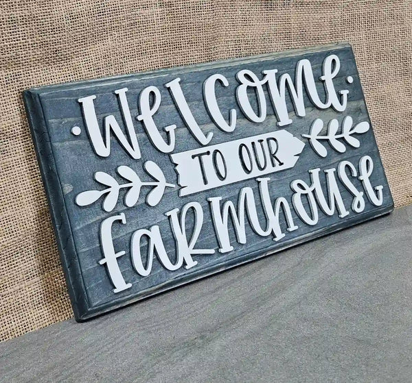 Angled view of a rectangular wooden sign with a blue stained finish and raised white script lettering that reads 'Welcome' at the top and 'farmhouse' at the bottom, with a white arrow pointing right that says 'TO OUR' in smaller black letters, flanked by white leafy branches, against a burlap background.