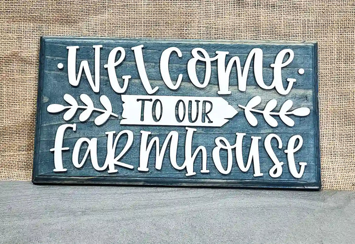 Front view of a rectangular wooden sign with a blue stained finish and raised white script lettering that reads 'Welcome' at the top and 'farmhouse' at the bottom, with a white arrow pointing right that says 'TO OUR' in smaller black letters, flanked by white leafy branches, against a burlap background.
