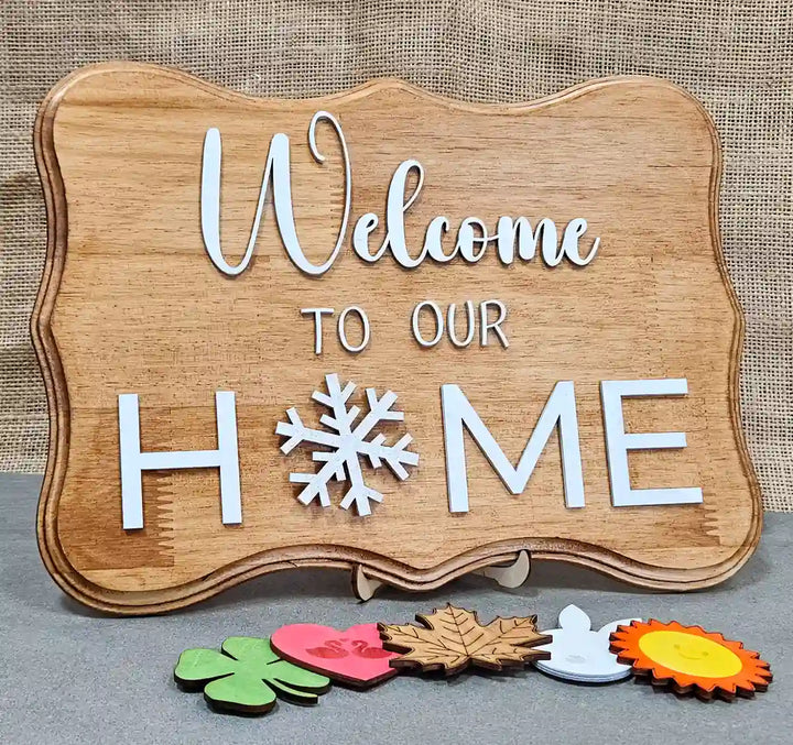 Wooden sign with 'Welcome' in white script, 'TO OUR' in smaller white block letters, and 'H❄ME' in larger white block letters with a snowflake replacing the 'O', displayed with six interchangeable magnetic shapes below: a clover, a heart with swans, a maple leaf, a bunny head, a sun, and a snowflake, against a burlap background.
