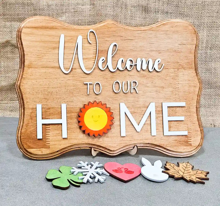 Wooden sign with 'Welcome' in white script, 'TO OUR' in smaller white block letters, and 'H☀️ME' in larger white block letters with a smiling sun replacing the 'O', displayed with six interchangeable magnetic shapes below: a clover, a snowflake, a heart with swans, a bunny, a maple leaf, and a sun, against a burlap background.