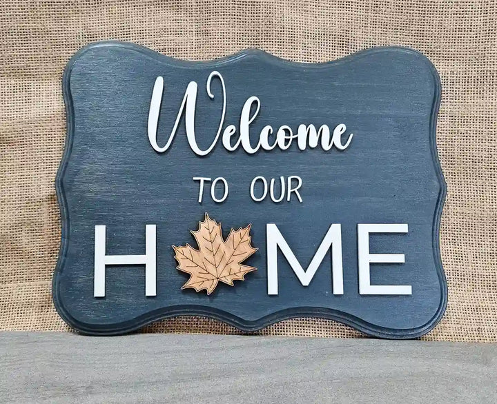 Wooden sign with a dark blue-washed background and 'Welcome' in white script, 'TO OUR' in smaller white block letters, and 'H🍁ME' in larger white block letters with a wooden maple leaf replacing the 'O', against a burlap background.