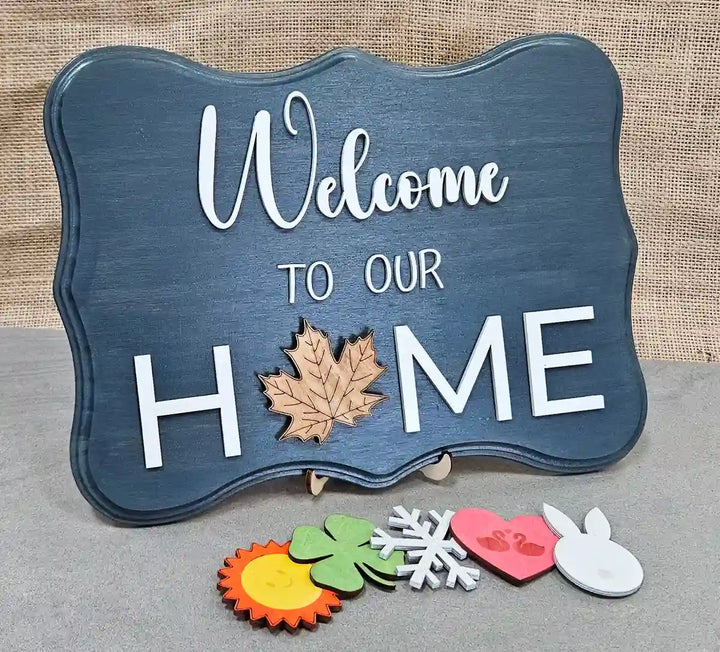 Wooden sign with a dark gray-washed background and 'Welcome' in white script, 'TO OUR' in smaller white block letters, and 'H🍁ME' in larger white block letters with a wooden maple leaf replacing the 'O', displayed with six interchangeable magnetic shapes below: a sun, a clover, a snowflake, a heart with swans, and a bunny, against a burlap background.