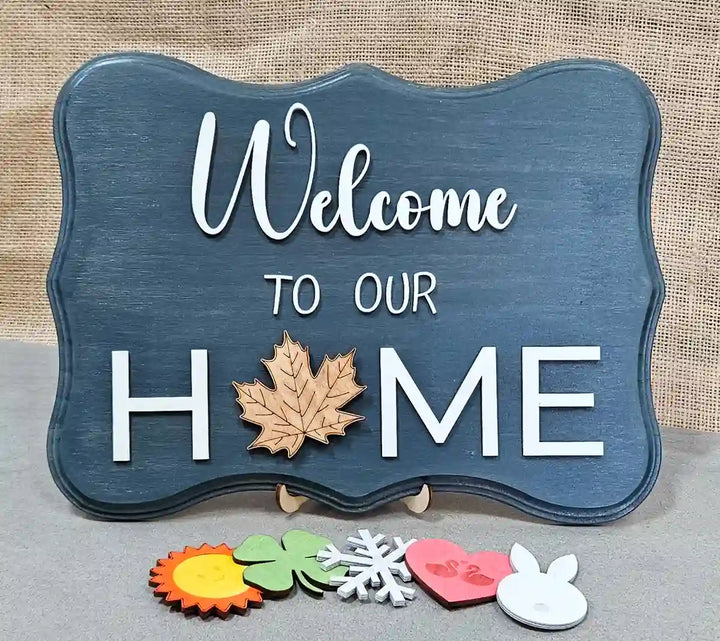 Wooden sign with a dark blue-washed background and 'Welcome' in white script, 'TO OUR' in smaller white block letters, and 'H🍁ME' in larger white block letters with a wooden maple leaf replacing the 'O', displayed with six interchangeable magnetic shapes below: a sun, a clover, a snowflake, a heart with swans, and a bunny, against a burlap background.