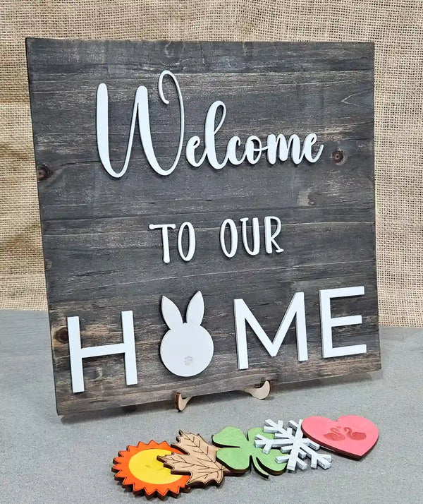 Wooden sign with a dark gray stained background and 'Welcome' in white script, 'TO OUR' in smaller white block letters, and 'H🐰ME' in larger white block letters with a white bunny silhouette replacing the 'O', displayed with five interchangeable magnetic shapes below: a sun, a maple leaf, a clover, a snowflake, and a heart with swans, against a burlap background.